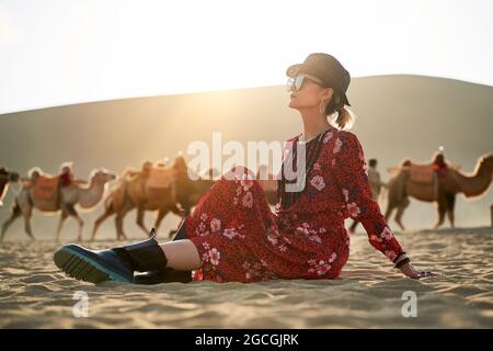 asiatische Frau in rotem Kleid sitzt in der Wüste Blick mit Karawane von Kamelen und riesige Sanddüne im Hintergrund Stockfoto