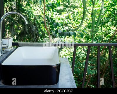 Edelstahl Wasserhahn mit Kurvenform und schwarz-weißen Keramikwaschbecken auf der Terrasse auf grünen Wald Natur Hintergrund. Stockfoto