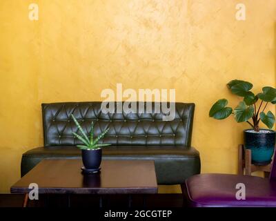 Dunkelgrüner Vintage Leder-Sofa-Sitz mit Stiften und Knöpfen neben Holztisch mit Pflanzentopf Dekoration auf gelbem Wandhintergrund im Wohnzimmer wi Stockfoto