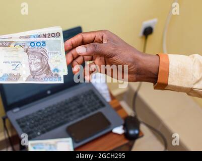 Eine afrikanische Hand, die nigrische Naira-Scheine, Bargeld oder Währung mit einem Laptop im Hintergrund erhält Stockfoto