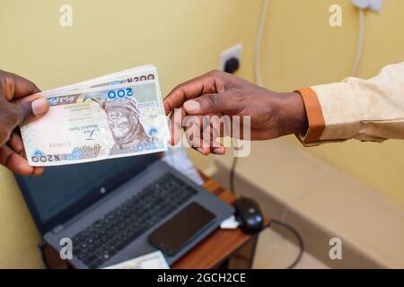 Eine afrikanische Hand, die nigrische Naira-Scheine, Bargeld oder Währung mit einem Laptop im Hintergrund erhält Stockfoto