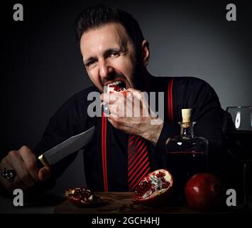 Erwachsene Mann in schwarzem Hemd und Hosenträger am Tisch serviert mit einer Flasche und einem Glas frischen Saft und beißt Granatapfel Stockfoto