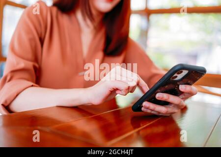 Nahaufnahme einer jungen schönen Frau, die auf dem Smartphone SMS schreibt und Apps und soziale Medien durchsucht, während sie im modernen Café auf Kaffee wartet Stockfoto
