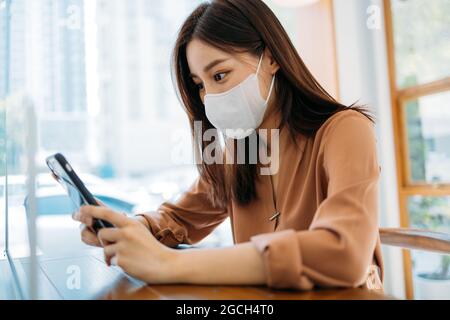 Schöne junge asiatische Frau sitzt in modernen Café tragen covid-19 Gesichtsschutz Maske mit und Surfen durch Smartphone während der Entspannung Stockfoto