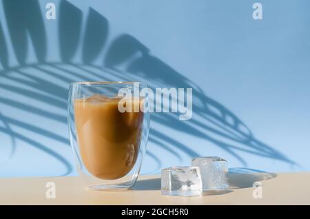 Iced Latte Coffee mit Eiswürfel auf dem Boden und Schatten von Kokosnussblatt auf braunem und blauem Hintergrund. Sommerdrink-Konzept. Stockfoto