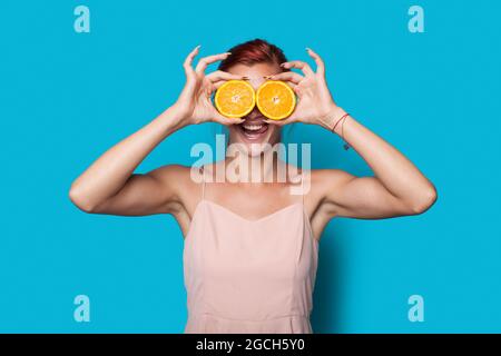 Schöne rothaarige Frau bedeckt ihr Auge mit geschnittenen Zitronen, die auf einer blauen Studiowand posieren Stockfoto