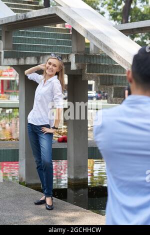 Schöne lächelnde junge Frau in weißer Sorte und Jeans posiert, als ihr Freund sie auf einer Digitalkamera fotografiert Stockfoto