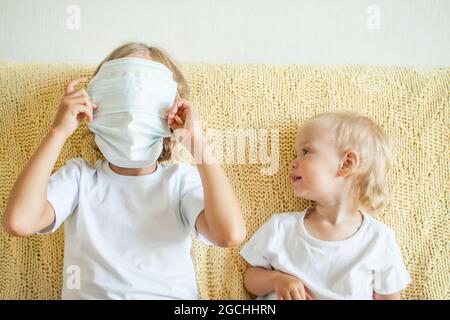 Portrait von zwei Schwestern, die medizinische Schutzmasken aufsetzen. Die ältere Schwester hilft der Jüngeren. Schwestern spielen Spaß. Coronavirus-Schutzkonzept Stockfoto