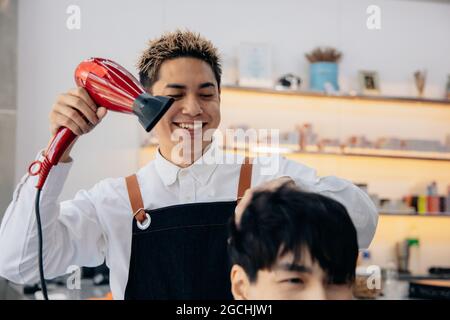 Porträt eines männlichen asiatischen Friseurs, der eine Schürze trägt und konzentriert arbeitet, während er in einem modernen Friseurladen die Haare eines hübschen jungen Mannes trocknet Stockfoto