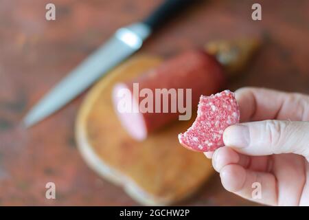 Ein unfokusster Schuss einer Hand, die ein Stück Salami über den Küchentisch hält. Ein Mann hält ein Stück Wurst in der Hand. Halbe Wurststange, Messer und Schnitt Stockfoto