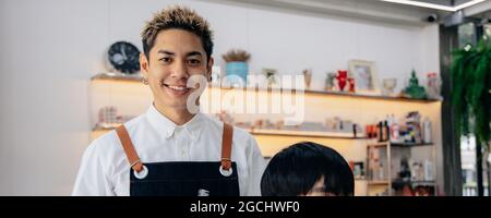 Porträt des erfolgreichen jungen asiatischen männlichen Friseur lächelnd trägt Schürze in modernen Salon hält Bürste und Schere bereit, Haare des Kunden schneiden Stockfoto