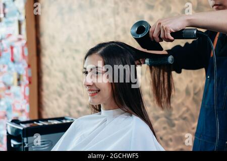 Porträt eines männlichen asiatischen Friseurs, der eine Schürze trägt und konzentriert arbeitet, während er in einem modernen Friseurladen hübsche junge Frauenhaare trocknet Stockfoto