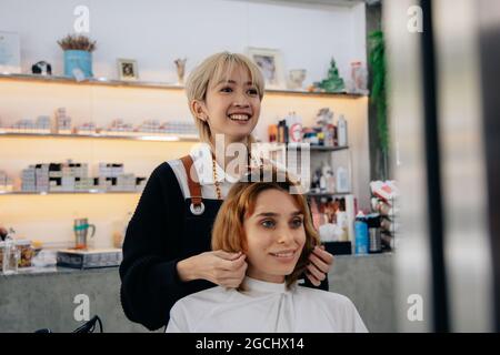 Porträt einer glücklichen jungen asiatischen Frau Friseurin in Schürze, während Friseursalon und legte Pflege Behandlung beim Styling lockiges Haar für Client Stockfoto