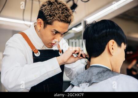 Glücklich junge asiatische männliche Friseur lächelnd trägt Schürze in modernen Salon hält Pinsel und Schere Schneiden Haar des Kunden Stockfoto