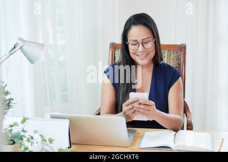 Eine positive, elegante Geschäftsfrau, die mit Kollegen über einen Messenger auf ihrem Smartphone kommuniziert, wenn sie am Schreibtisch arbeitet Stockfoto