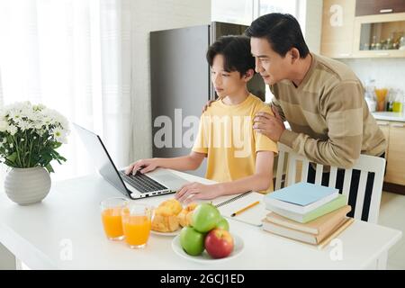 Stolzer Vater hilft seinem Sohn im Teenageralter bei der Hausaufgabe auf dem Laptop, wenn er aufgrund einer Pandemie online studiert Stockfoto