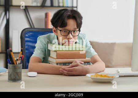 Porträt eines nachdenklichen Jungen im Teenageralter, der sich auf einen Bücherstapel auf seinem Schreibtisch lehnt und die Kamera anschaut Stockfoto