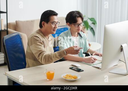 Ein reifer Mann hilft seinem Sohn im Teenageralter bei Hausaufgaben, wenn er am Schreibtisch sitzt und auf den Computerbildschirm schaut Stockfoto