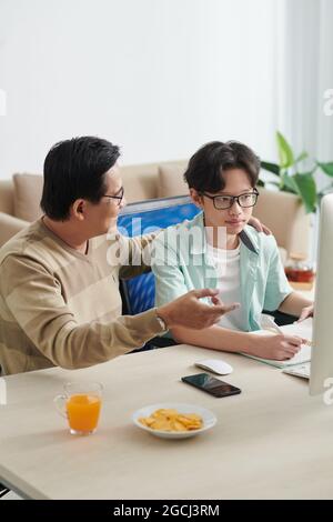 Lächelnder Vater umarmte seinen Sohn und erklärte sein schwierigst mathematisches Thema auf dem Computerbildschirm Stockfoto