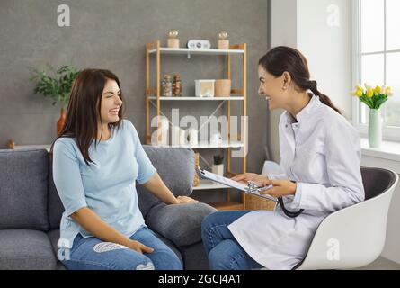 Glückliche junge Ärztin und Patientin, die während eines Hausbesuches redet und lacht Stockfoto