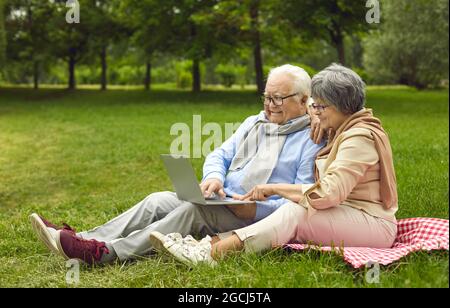 Glückliches Seniorenpaar, das einen Laptop benutzt, während es zusammen auf einer Parkwiese sitzt Stockfoto