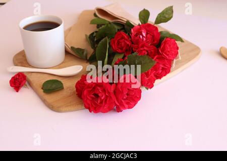 Tasse Kaffee umgeben von roten Rosen auf dem hellrosa Hintergrund Stockfoto