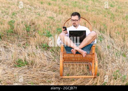 Junger Geschäftsmann, der sein Smartphone hält, während er in einem Schaukelstuhl am Computer arbeitet. Erwachsener downshifter arbeitet an einem Laptop in der Mitte eines f Stockfoto