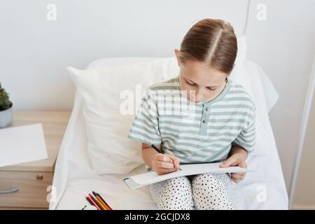 Minimales Porträt eines kleinen Mädchens im Krankenhauszimmer, das Bilder zeichnet, während es auf dem Bett sitzt, Raum kopieren Stockfoto