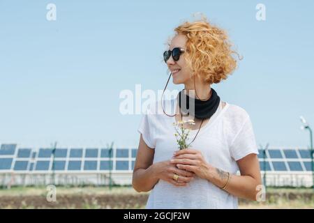 Lächelnde Frau, die die Aussicht genießt und Kamillen hält. Über dem Himmel, niedriger Winkel. Elektrische Sonnenkollektoren in einer Entfernung. Stockfoto