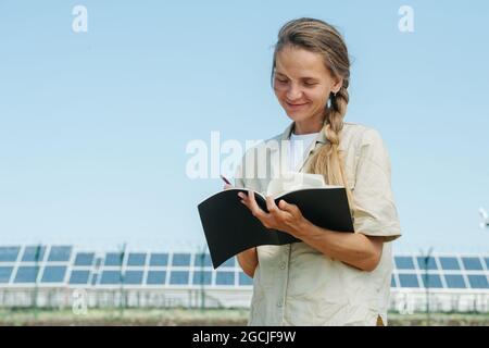 Fröhliche lächelnde Frau, die Dinge auf ihren Notizblock schrieb. Über dem Himmel, niedriger Winkel. Elektrische Sonnenkollektoren in einer Entfernung. Stockfoto