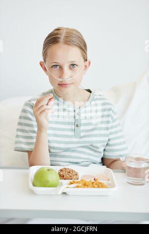 Minimales Porträt eines Mädchens, das im weißen Krankenhauszimmer gesundes Mittagessen isst und die Kamera anschaut Stockfoto