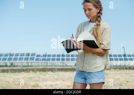 Beschäftigte Frau, die Dinge auf ihren Notizblock schreibt. Über dem Himmel, niedriger Winkel. Elektrische Sonnenkollektoren in einer Entfernung. Stockfoto