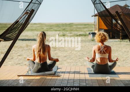 Zwei Frauen meditieren von hinten vor der Türschwelle eines großen Zeltes. Sitzen in locker Pose, Yoga machen. Wunderschöne, schlichte Steppe rundherum. Anti-Moskitonetz Stockfoto