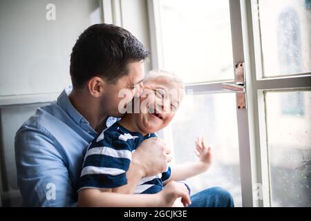 Vater mit Happy Down-Syndrom Sohn in Innenräumen zu Hause, küsst seine Wange. Stockfoto