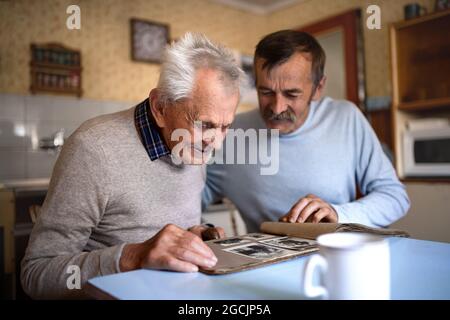 Porträt eines Mannes mit einem älteren Vater, der zuhause am Tisch sitzt und ein Fotoalbum anschaut. Stockfoto