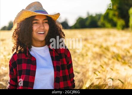 Schöne glücklich gemischte Rasse afroamerikanische biracial weibliche Mädchen Teenager junge Frau mit perfekten Zähnen trägt Cowboy Hut und kariertes Hemd sitzen in Stockfoto