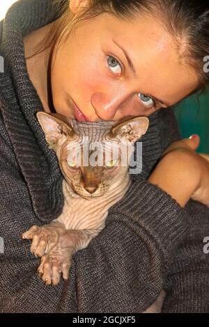 Ein schönes, braunes, grauäugiges Mädchen umarmt die Lieblingskatze der kahlen, grünäugigen Kanadierin Sphynx. Porträt einer Sphinx Kitty und Herrin, junge Frau. World Cat Da Stockfoto