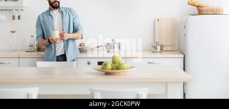 Entspannen Sie sich zu Hause. Junger unkennbarer Mann trinkt Morgenkaffee in der Küche und träumt, Panorama mit leerem Raum Stockfoto