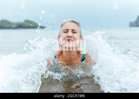 Aktive ältere grauhaarige Frau, die im Meerwasser schwimmt, während sie den Sommer genießt und an der Küste einen gesunden Lebensstil praktiziert Stockfoto