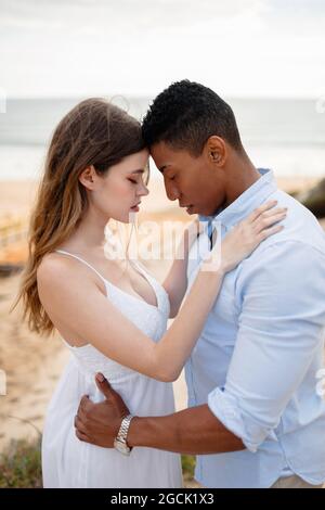 Seitenansicht eines liebevollen multirassischen Brautpaares, das sich am Hochzeitstag auf einem sandigen Hügel vor dem Hintergrund des Meeres umarmt Stockfoto