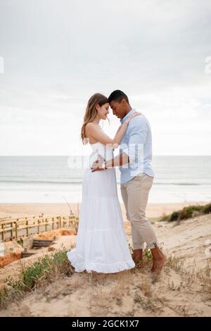 Seitenansicht eines liebevollen multirassischen Brautpaares, das sich am Hochzeitstag auf einem sandigen Hügel vor dem Hintergrund des Meeres umarmt Stockfoto