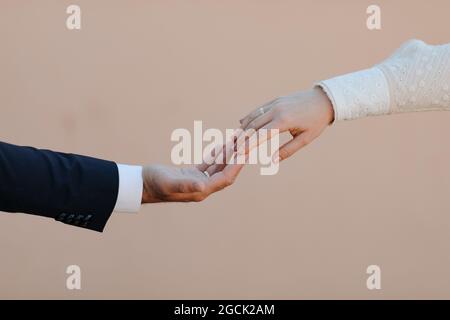 Crop anonyme romantische frisch verheiratetes Paar in eleganten stilvollen Hochzeitskleidung mit Verlobungsringen berühren Hände sanft vor beigem Hintergrund Stockfoto