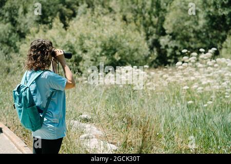 Seitenansicht einer nicht erkennbaren Freiwilligen-Frau in blauem Hemd mit Rucksack, die während der Öko-Kampagne im Sommerwald die Natur mit einem Fernglas erkundet Stockfoto