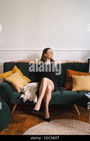 Selbstbewusste Geschäftsfrau mit Brille in einem schwarzen klassischen Kleid, die in einem modernen Apartment auf einer Couch sitzt. Grünes Plüschsofa mit gelben Kissen Stockfoto