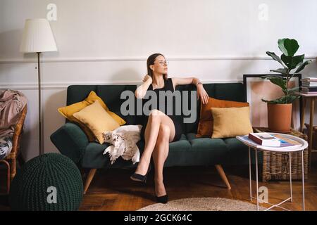 Selbstbewusste Geschäftsfrau mit Brille in einem schwarzen klassischen Kleid, die in einem modernen Apartment auf einer Couch sitzt. Grünes Plüschsofa mit gelben Kissen Stockfoto