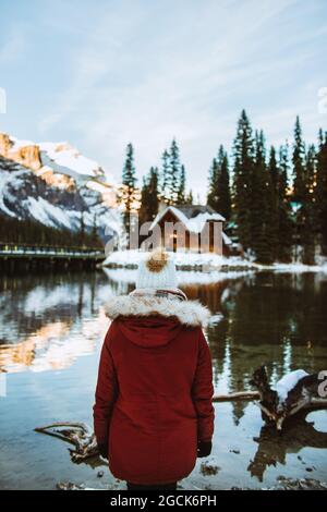 Rückansicht einer nicht erkennbaren weiblichen Touristin in Oberbekleidung, die Holzhütte und Bergrücken bewundert, während sie auf der verschneiten Küste des Emerald Lake auf Wint steht Stockfoto