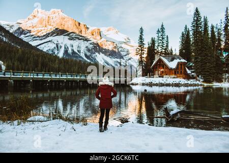 Rückansicht einer nicht erkennbaren weiblichen Touristin in Oberbekleidung, die Hände in den Taschen hält und während eines Spazierganges auf verschneiten Bergen die Holzhütte und den Bergkamm bewundert Stockfoto