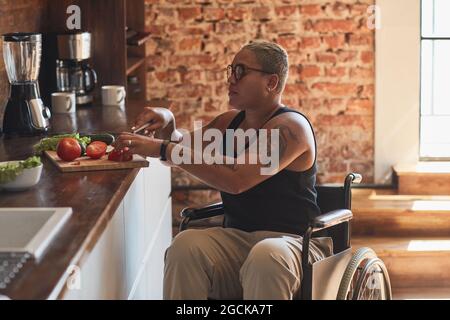 Seitenansicht Porträt einer zeitgenössischen tätowierten Frau mit Behinderung Kochen Abendessen zu Hause, Kopierraum Stockfoto