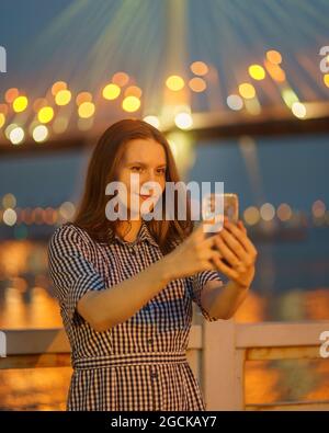 Junge Frau, die in der Nacht Selfie auf schönen verschwommenen Lichtern der Brücke fotografiert Stockfoto