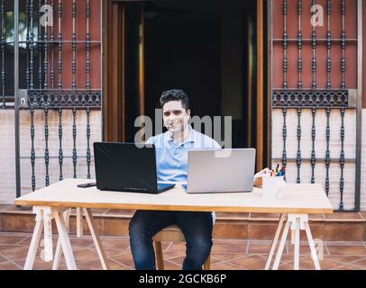 Lächelnder junger Mann, der mit Kryptowährungen handelt, sitzt auf einer Terrasse Stockfoto